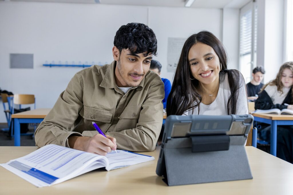 Foto einer Schülerin und einem Schüler, die gemeinsam mit Buch und Tablet im Klassenzimmer an den SABEL Schulen arbeiten. Sinnbild für die angedachten Wirtschaftsschule am Bildungscampus Taufkirchen.
